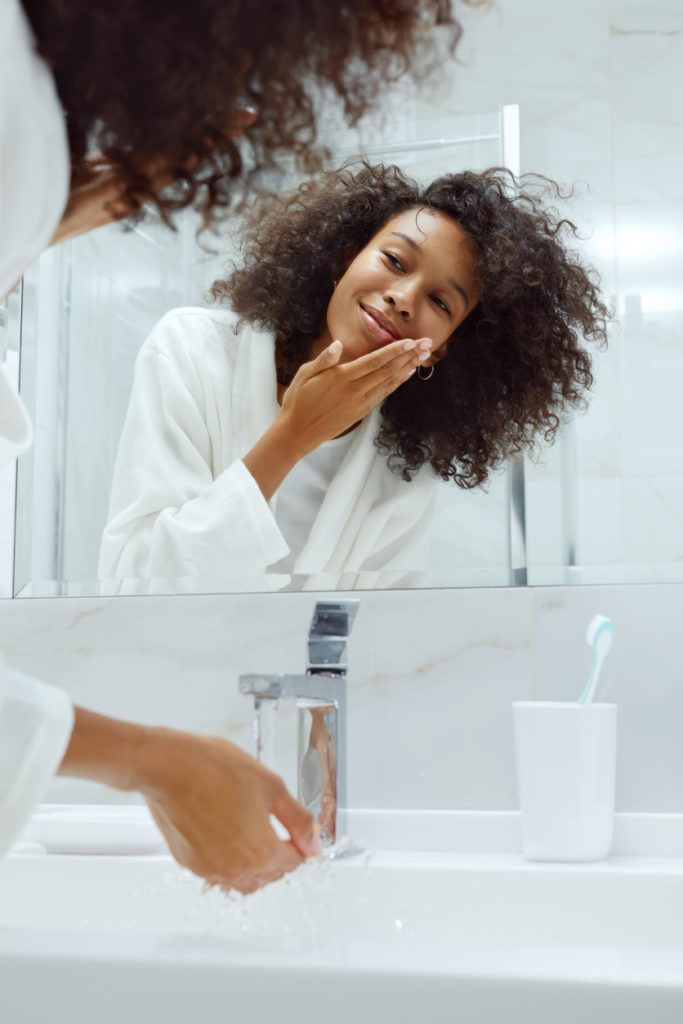 Skin care. Woman washing face with clean water in sink looking in mirror at bathroom. Portrait of beautiful smiling african girl with afro hair in white bath robe cleaning facial skin closeup