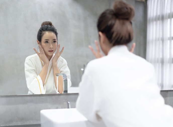 Facial Beauty.Young beautiful Asian woman applying moisturizer cream on her pretty face and looking to mirror at bathroom.Natural skin care and people Concept.